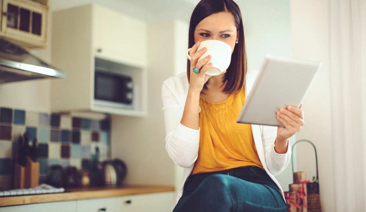 Young female sitting on a counter top drinking coffee while looking at her tablet. 