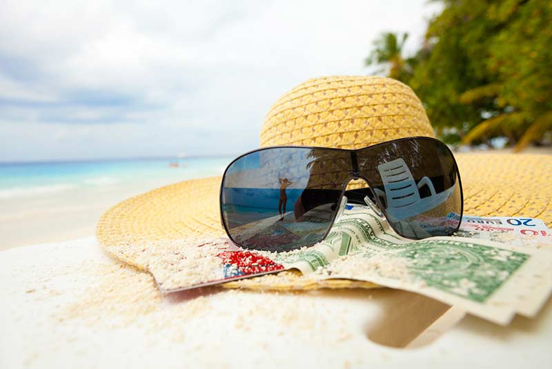 A beach hat, sunglasses, and money sitting on a sandy beach by the ocean.